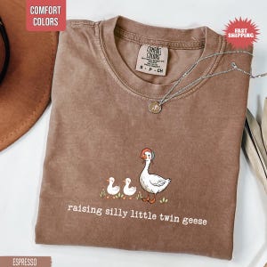 May include: Espresso-colored t-shirt with a crew neck and the text "raising silly little twin geese." The shirt features a graphic of a goose and two goslings. A silver necklace with a pendant is on the shirt.