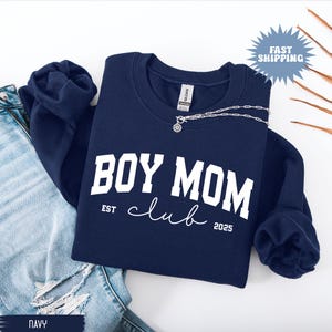 May include: Navy blue sweatshirt with the text "BOY MOM club" in white block letters and cursive. The sweatshirt also has the text "EST 2025". A silver necklace with a pendant is on the sweatshirt.
