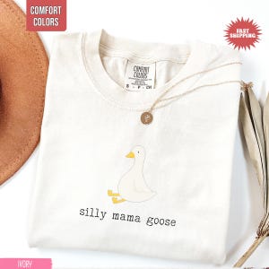 May include: Ivory-colored t-shirt with a cartoon goose illustration and the text "silly mama goose." The shirt has a crew neck and short sleeves. A gold necklace with a pendant is draped on the shirt.