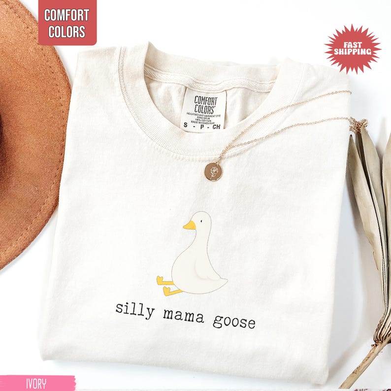 May include: Ivory-colored t-shirt with a cartoon goose illustration and the text "silly mama goose." The shirt has a crew neck and short sleeves. A gold necklace with a pendant is draped on the shirt.