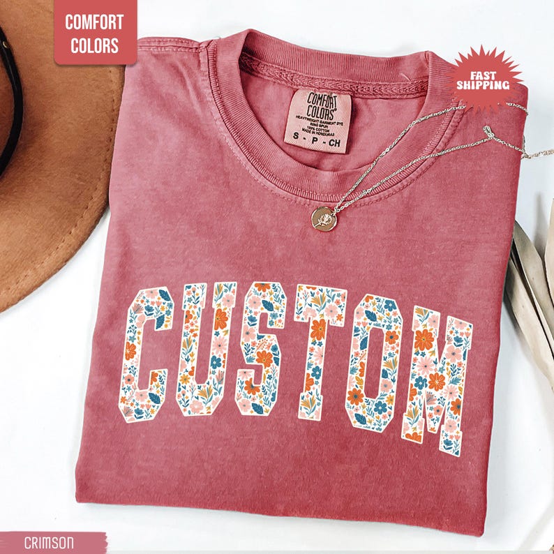 K&ouml;nnte beinhalten: Karmesinrotes T-Shirt mit dem Wort "CUSTOM" in wei&szlig;en Blockbuchstaben, gef&uuml;llt mit einem floralen Muster. Das Shirt hat einen Rundhalsausschnitt und kurze &Auml;rmel. Eine silberne Halskette mit Anh&auml;nger liegt &uuml;ber dem Shirt.