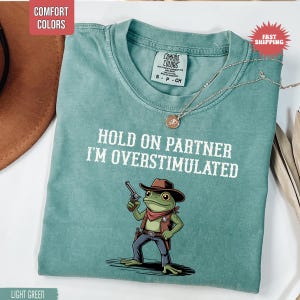 May include: Light green t-shirt with the text "HOLD ON PARTNER I'M OVERSTIMULATED" and a cartoon frog wearing a cowboy hat and holding a gun. The shirt has a crew neck and short sleeves. A silver necklace with a pendant is on the shirt.