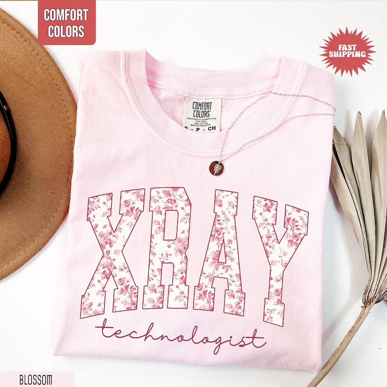 May include: A light pink t-shirt with the words "XRAY" and "technologist" in a floral design. The shirt has a crew neck and short sleeves. A gold necklace with a pendant is draped across the shirt.
