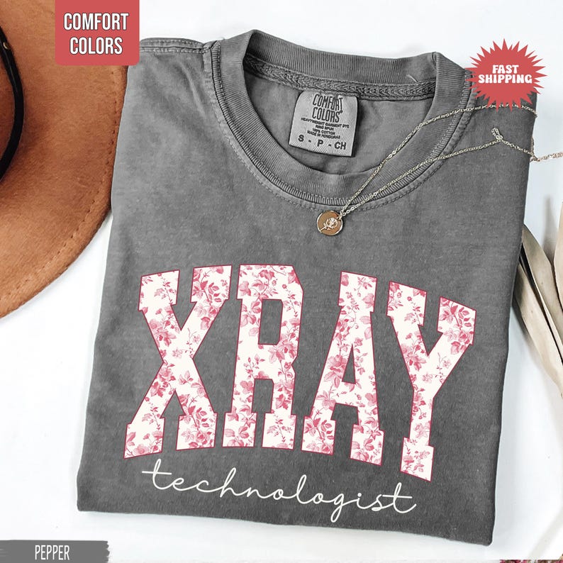 May include: A gray Comfort Colors t-shirt with the word "XRAY" in large, floral-patterned letters, and the word "technologist" below. A silver necklace with a pendant rests on the shirt.
