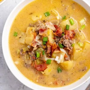 May include: A close-up shot of a bowl filled with creamy potato soup. The soup is a light yellow color and contains chunks of potatoes, bacon, ground meat, and green onions. A second bowl of soup is partially visible in the upper left corner.