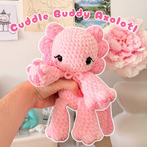 Könnte beinhalten: Ein hellrosa gehäkeltes Axolotl-Stofftier mit weicher, strukturierter Garnoberfläche, großen schwarzen Augen mit gestickten Wimpern und einer kleinen Schleife am Hals. Der Text Cuddle Buddy Axolotl! ist oben im Bild zu sehen.