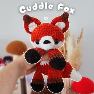 May include: A handmade crochet fox toy in shades of orange, brown, black, and white. The fox has large ears, a white muzzle, and black eyes. The text "Cuddle Fox" is above the fox.