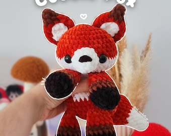 May include: A handmade crochet fox toy in shades of orange, brown, black, and white. The fox has large ears, a white muzzle, and black eyes. The text "Cuddle Fox" is above the fox.