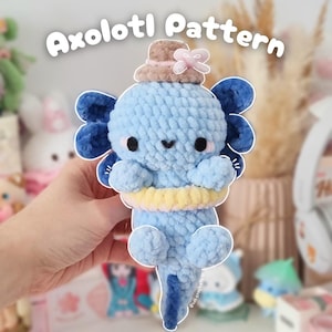 May include: A blue crocheted axolotl wearing a brown hat with a pink bow and a yellow and pink life preserver. The axolotl has black eyes and a small smile. The text "Axolotl Pattern" is written above the axolotl.