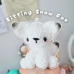 Puede incluir: Un zorro de nieve de ganchillo blanco hecho a mano con detalles en negro y gris en las orejas. El zorro tiene una nariz y ojos negros, con mejillas rosadas. El texto "Sitting Snow Fox" está arriba.
