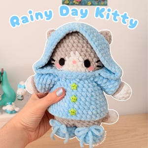 May include: A handmade crochet cat plushie wearing a light blue hooded jacket with star-shaped buttons. The plushie has gray and white fur, black eyes, and pink cheeks. The text "Rainy Day Kitty" is above the plushie.