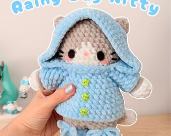 May include: A handmade crochet cat plushie wearing a light blue hooded jacket with star-shaped buttons. The plushie has gray and white fur, black eyes, and pink cheeks. The text "Rainy Day Kitty" is above the plushie.