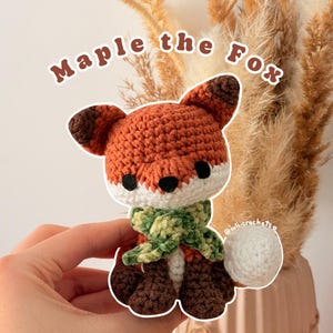 Puede incluir: Un pequeño zorro de ganchillo llamado Maple the Fox hecho con hilo naranja, blanco y marrón oscuro. El zorro tiene ojos negros, una nariz marrón oscura y una bufanda de hilo verde jaspeado. El texto Maple the Fox se ve sobre la figura.