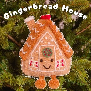 May include: A plush gingerbread house ornament with the text "Gingerbread House". The ornament is tan with white icing details, red accents, and a smiling face. It has two orange feet and is set against a green, leafy background.