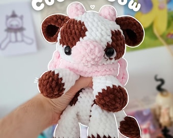 May include: A handmade plush cow toy with a white, brown, and pink color scheme. The toy has large black eyes and a pink scarf. The text "Cuddle Cow" is above the toy. The toy is being held by a person.