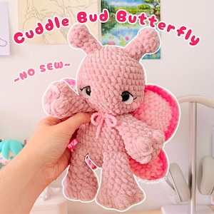 May include: A pink crochet plush butterfly toy with a textured yarn finish, large black eyes, and a small bow at the neck. The text Cuddle Bud Butterfly and ~NO SEW~ are visible in the image. The plush features rounded wings with pink accents.