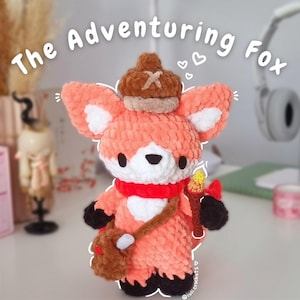 May include: A crocheted fox plush toy wearing a brown hat, a red scarf, and a brown satchel. The fox is holding a stick with a yellow flame on top. The text "The Adventuring Fox" is written above the fox.