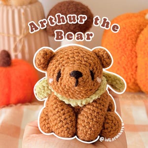 May include: A small brown crocheted bear toy with a light green textured scarf around its neck. The bear has black embroidered eyes and a black nose, with small pink blush marks on its cheeks. The text Arthur the Bear is visible above it.