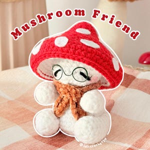 May include: A crocheted mushroom plush toy with a red cap featuring white felt spots, a white body, and a brown textured scarf. The figure wears thin, round, dark-rimmed glasses and has closed eyes. The text "Mushroom Friend" appears above.