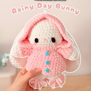 Puede incluir: Un peluche de conejo tejido a ganchillo de color blanco que lleva un abrigo rosa con capucha y tres botones azul claro en forma de estrella. El conejo tiene ojos negros, mejillas rosadas y orejas largas. El texto Rainy Day Bunny aparece arriba.
