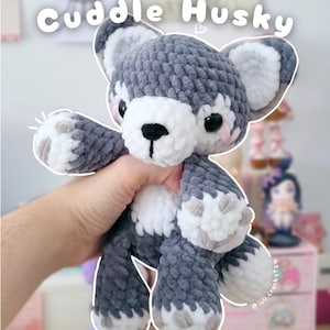 May include: A handmade crochet husky dog plushie in grey, white, and black. The plush toy has a fluffy texture, with the text "Cuddle Husky" above it. The husky is being held by a person.