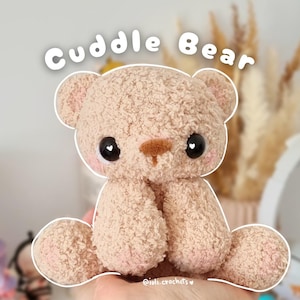 Könnte beinhalten: Ein handgemachter, hellbrauner Teddybär mit dem Text "Cuddle Bear". Der Bär hat große schwarze Augen mit weißen herzförmigen Glanzlichtern, eine braune Nase und rosige Wangen. Das Fell des Bären ist strukturiert und sieht weich aus, mit einem niedlichen, freundlichen Ausdruck.