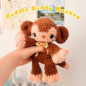 May include: A brown and light pink crocheted monkey plush toy with large dark eyes and a yellow ribbon tied around its neck. The text Cuddle Buddy Monkey is displayed in yellow letters above the plush, which has a soft, textured yarn finish.