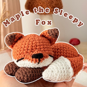 Peut inclure: Une petite figurine de renard crochetée à la main en position endormie, avec du fil orange, des pattes et des pointes d'oreilles marron foncé, et une queue à bout blanc. Le texte "Maple the Sleepy Fox" est affiché au-dessus en blanc.