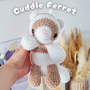 Könnte beinhalten: Ein handgefertigtes gehäkeltes Frettchen-Plüschtier in Weiß, Braun und Rosa. Das Frettchen hat einen freundlichen Ausdruck und hebt eine Pfote. Der Text "Cuddle Ferret" steht über dem Spielzeug. Das Spielzeug ist etwa 15 cm groß.