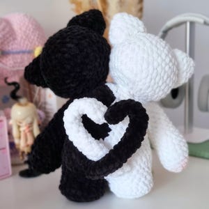 Puede incluir: Dos juguetes de peluche de crochet, uno negro y otro blanco, est&aacute;n de pie uno al lado del otro. Sus colas est&aacute;n entrelazadas para formar una forma de coraz&oacute;n.