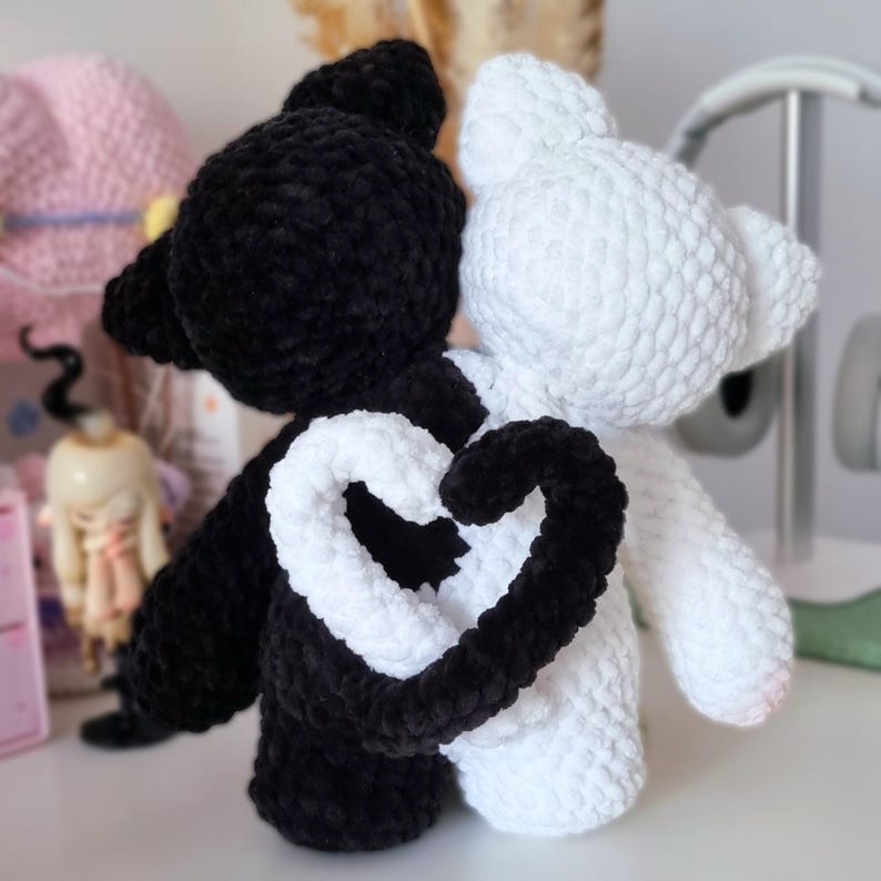 Puede incluir: Dos juguetes de peluche de crochet, uno negro y otro blanco, est&aacute;n de pie uno al lado del otro. Sus colas est&aacute;n entrelazadas para formar una forma de coraz&oacute;n.