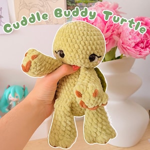 May include: A light green crochet turtle plush toy with a textured yarn finish, dark brown accents on the feet, and large black eyes with eyelashes. The text Cuddle Buddy Turtle is displayed in green letters above the plush turtle figure.