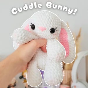Könnte beinhalten: Ein weißer Häkelhase mit rosa Akzenten an Ohren und Wangen wird in einer Hand gehalten. Der Hase hat schwarze Augen und der Text "Cuddle Bunny!" steht darüber. Es ist ein weiches, plüschiges Spielzeug.
