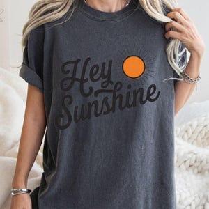 Op de afbeelding: Een antracietgrijs T-shirt met korte mouwen en de zwarte cursieve tekst Hey Sunshine op de voorzijde. Rechts van de tekst staat een afbeelding van een feloranje zon met dunne zwarte lijnen die vanuit het midden naar buiten stralen.