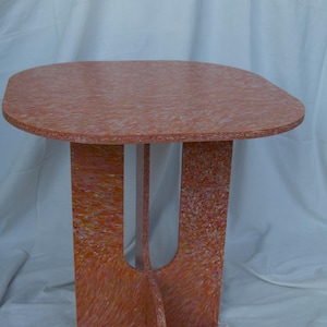 May include: A small, modern side table with a rounded rectangular top and a unique base. The table is made of a composite material with a speckled orange and pink pattern. The table's dimensions are approximately 61 cm long, 46 cm wide, and 51 cm high.