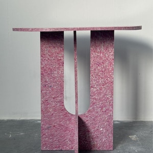 May include: A modern side table with a unique design. The table is constructed from a speckled pink material, featuring a rectangular top and a base with a U-shaped cutout. The table's dimensions are approximately 30 inches in height and 20 inches in width.