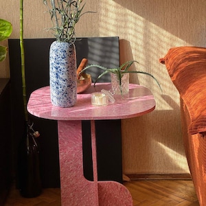 May include: A pink side table with a unique, asymmetrical design. The table has a round top and a patterned surface. A blue and white speckled vase holds orange flowers. Other items on the table include a small candle and a glass with a plant.