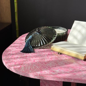 May include: A decorative glass jar with a lid and a blue tassel sits on a pink table. The jar is dark green and has a ribbed design. A small candle and a book are also on the table. The table has a unique speckled pattern.