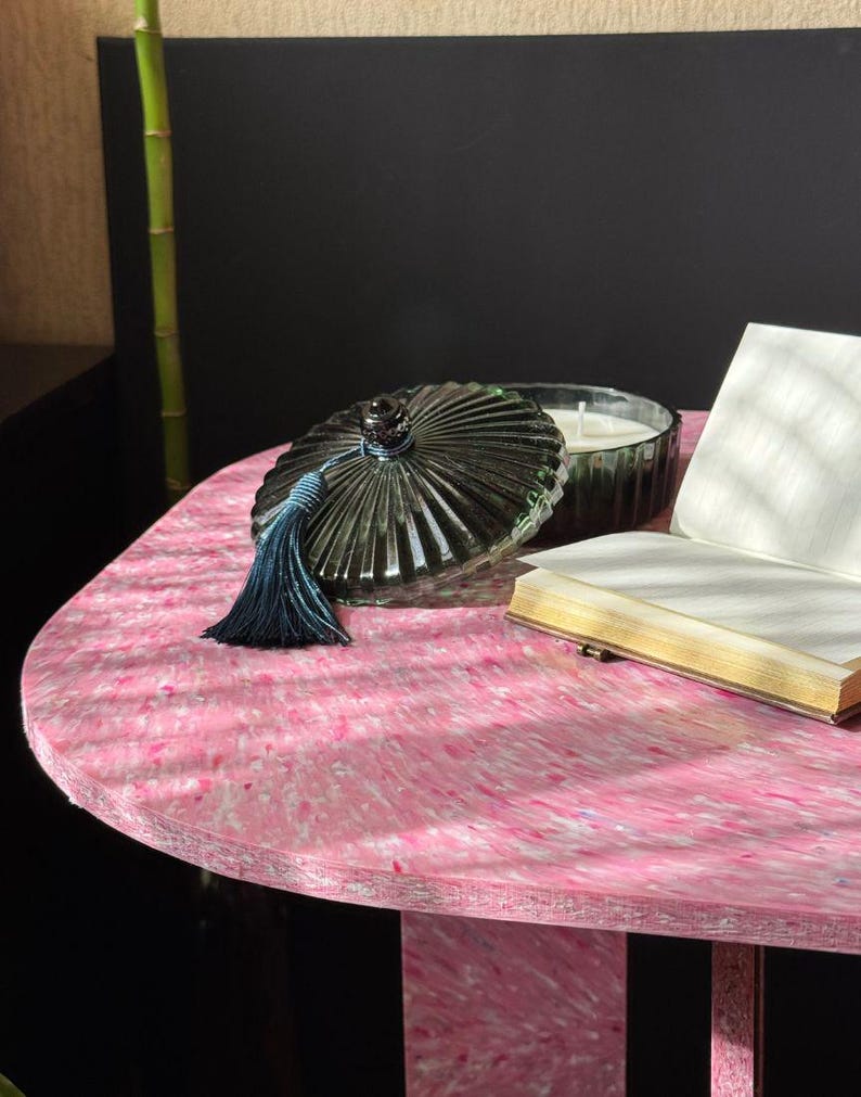 May include: A decorative glass jar with a lid and a blue tassel sits on a pink table. The jar is dark green and has a ribbed design. A small candle and a book are also on the table. The table has a unique speckled pattern.
