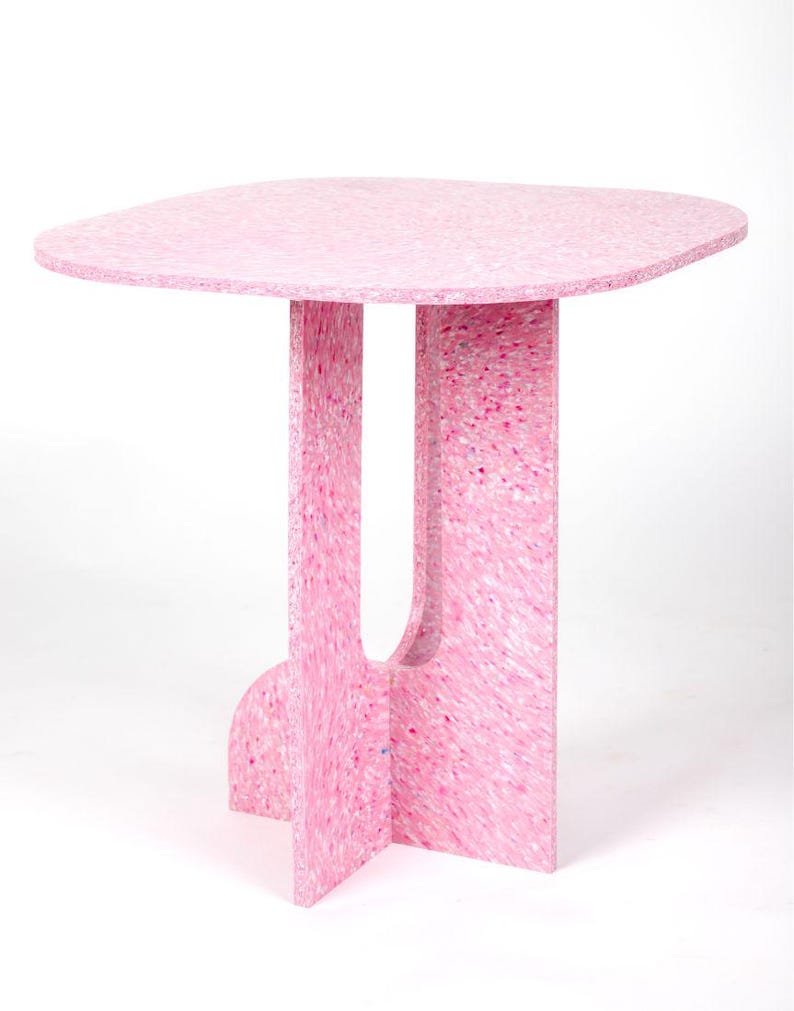 May include: A pink side table with a unique design. The table has a rounded, irregular top and a base with two vertical supports and a curved crossbar. The table is made of a speckled pink material, and measures approximately 28 inches high.