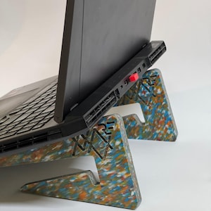 Könnte beinhalten: Ein Laptop-Computer steht auf einem bunten, recycelten Laptop-Ständer aus Kunststoff. Der Ständer hat ein einzigartiges Design mit einem Logo, das "XAR" lautet.