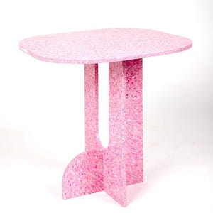 May include: A pink side table with a rounded rectangular top and a unique base design. The table is made of a speckled material, with shades of pink and white. The table is approximately 30 inches tall.