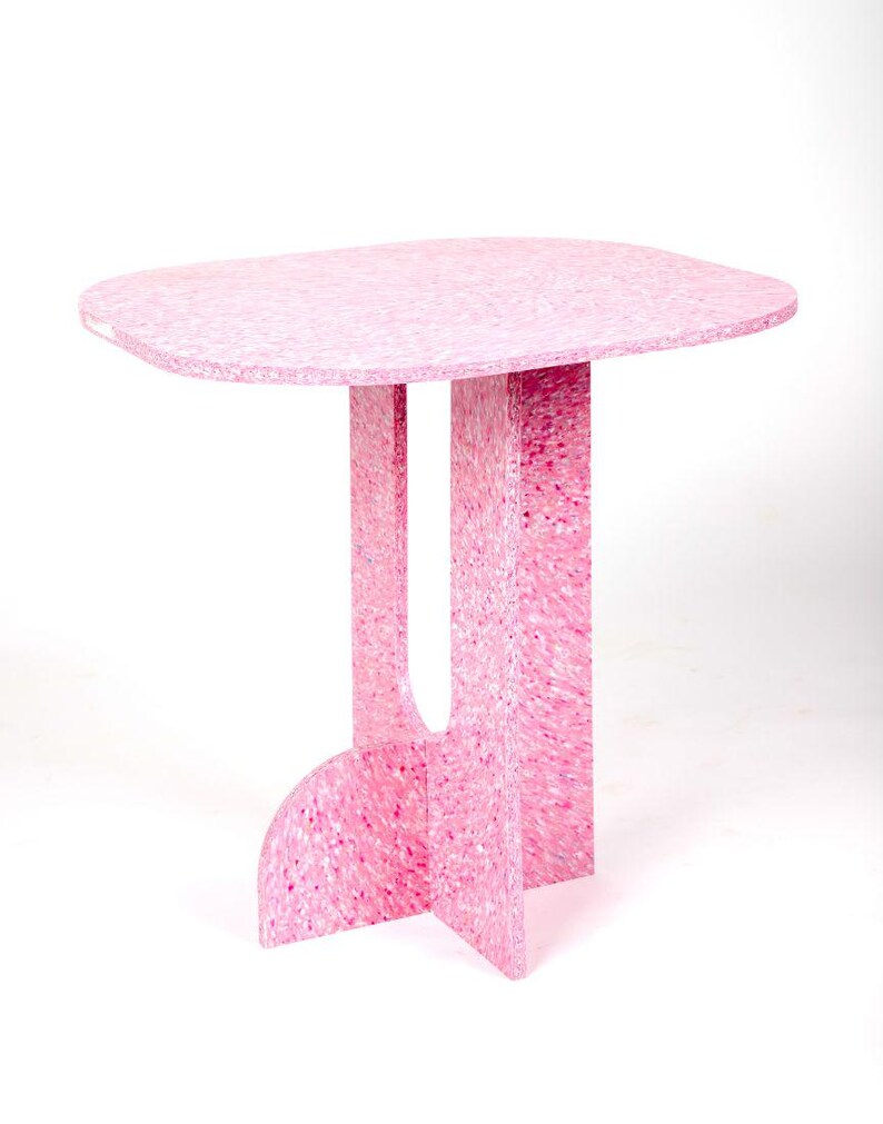 May include: A pink side table with a rounded rectangular top and a unique base design. The table is made of a speckled material, with shades of pink and white. The table is approximately 30 inches tall.