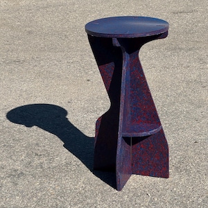 May include: A tall, wooden stool with a round top and a small shelf. The stool is painted in a speckled pattern of blue and red. The stool's design features a curved leg and a sturdy base.