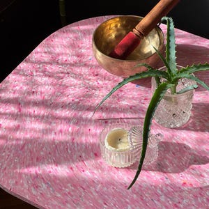 May include: A pink and white speckled table with a brass singing bowl and a wooden mallet. A small aloe vera plant in a glass container and a small candle in a glass jar are also on the table. The table has a unique, organic shape.