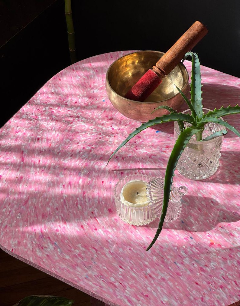 May include: A pink and white speckled table with a brass singing bowl and a wooden mallet. A small aloe vera plant in a glass container and a small candle in a glass jar are also on the table. The table has a unique, organic shape.
