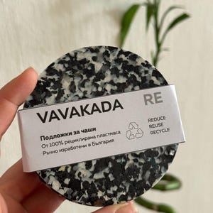 Può includere: Un sottobicchiere rotondo con un motivo maculato bianco e nero. Il sottobicchiere ha un'etichetta bianca con il testo "VAVAKADA RE" e testo in un'altra lingua. L'etichetta include anche le parole "REDUCE, REUSE, RECYCLE".