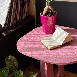 May include: A pink, rounded-edge table with a textured surface. A small, open book sits on the table, next to a bright pink flower pot holding a bonsai tree. A stack of vinyl records is visible in the background.