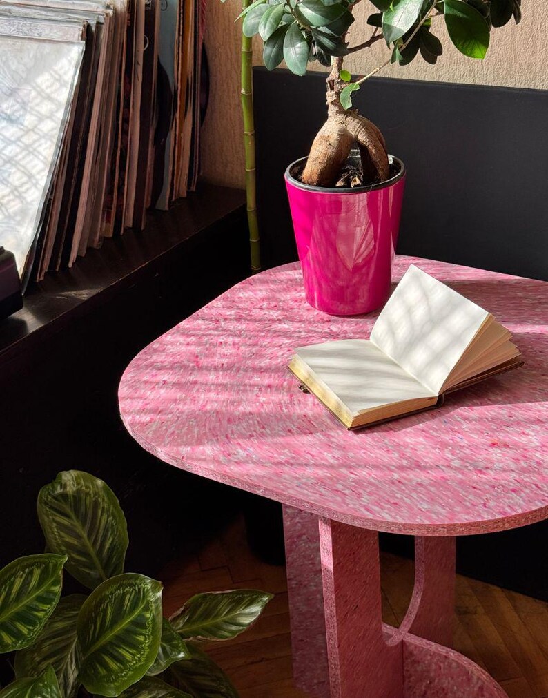 May include: A pink, rounded-edge table with a textured surface. A small, open book sits on the table, next to a bright pink flower pot holding a bonsai tree. A stack of vinyl records is visible in the background.