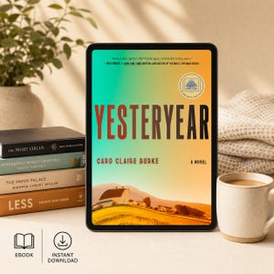 以下が含まれることがあります： デジタルタブレットには、Caro Claire Burke著の「Yesteryear」の表紙が表示されています。表紙は、グラデーションの背景を持つ納屋のイラストが特徴です。タブレットの横には、積み重ねられた本、コーヒーマグ、植物があります。